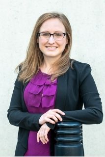 Kaitlyn Bean standing with her elbow resting on a black pole. She has long, light brown hair, is wearing dark rimmed glasses, and a black blazer and purple shirt.